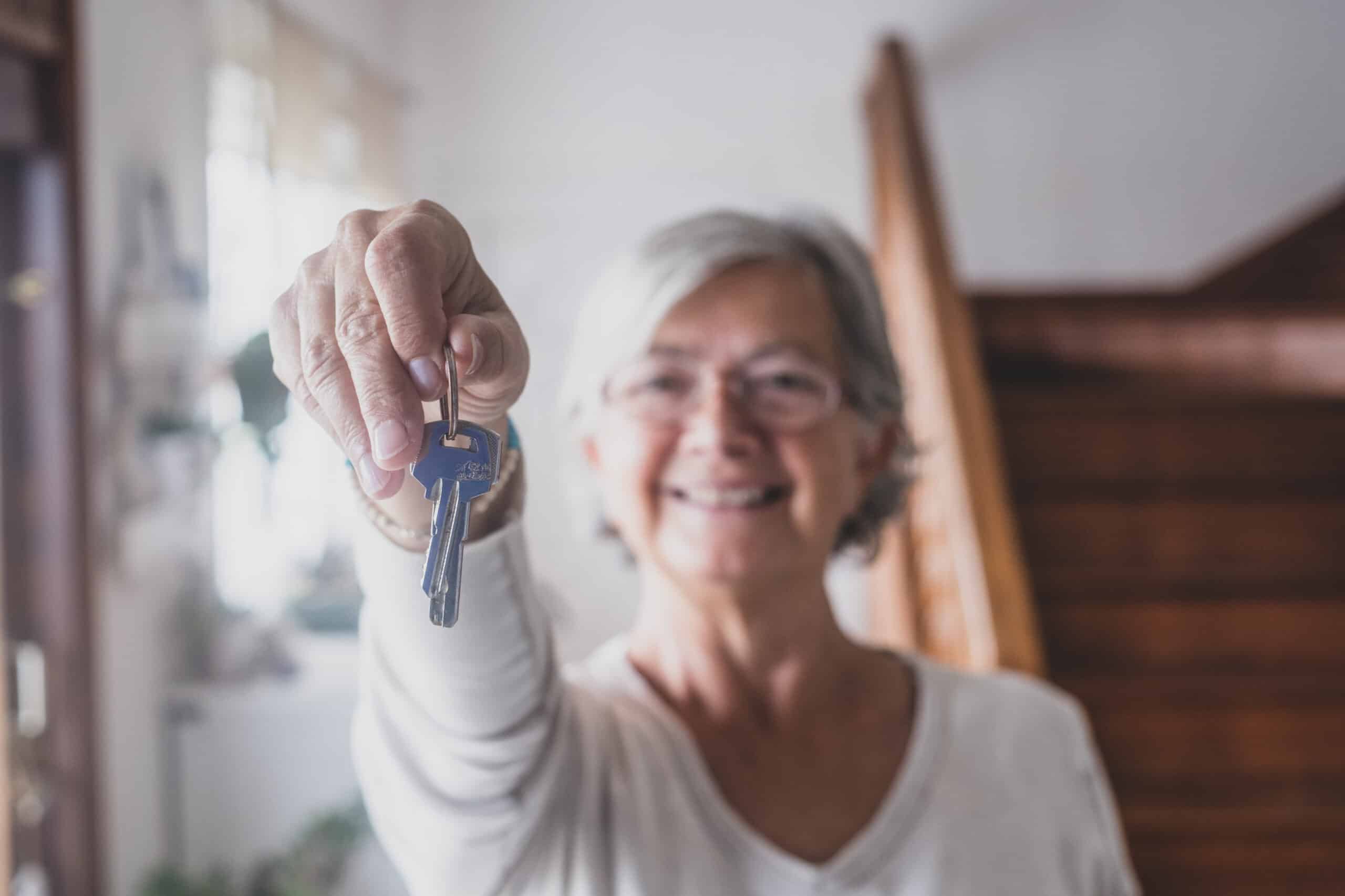 Happy senior old aged woman customer landlord hold key to new house apartment give to camera, older retired female hand real estate owner make sale purchase property deal concept, close up view